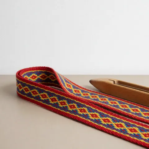 Ceinture tissée aux motifs géométriques rouges, jaunes et bleus posée sur une surface beige, accompagnée d'un outil en bois, idéale pour un atelier artisanal.