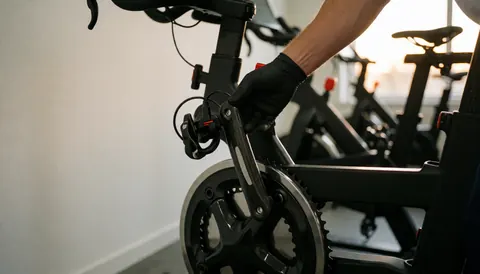 Main gantée de noir réglant un capteur de puissance sur un vélo d'appartement dans un gymnase moderne avec un mur blanc et une lumière naturelle.