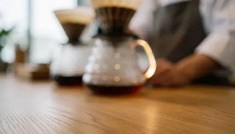 Carafe en verre remplie de café sur une table en bois, avec un filtre à café et une personne en arrière-plan, idéal pour les cafés.