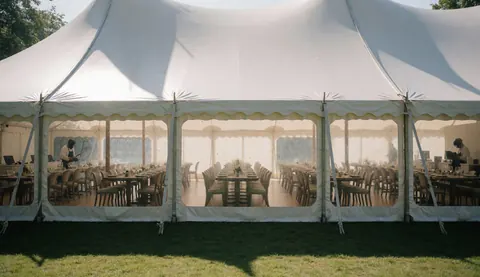 Tente blanche pour événement avec tables et chaises disposées, personnel travaillant à l'intérieur, lumière naturelle filtrée, jardin extérieur visible.