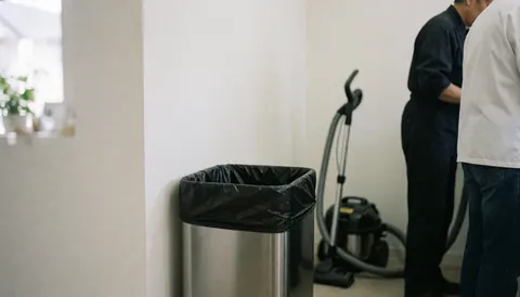 Poubelle avec sac noir dans une cuisine propre, aspiratrice et deux personnes travaillant en arrière-plan, murs blancs et lumière naturelle.