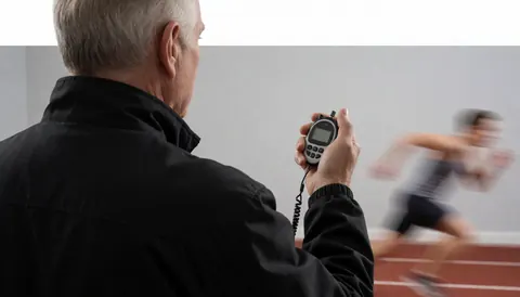 Entraîneur de football tenant un chronomètre numérique, observant un athlète courir sur une piste d'athlétisme, vu de dos, scène sportive réaliste.