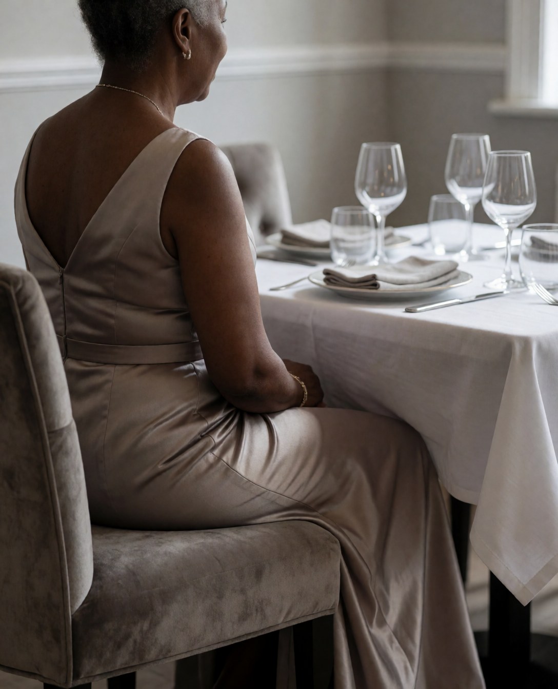 Une femme adulte en robe formelle est assise sur une chaise en velours, regardant une table élégante avec vaisselle et verres à vin dans un restaurant aux murs gris clair.