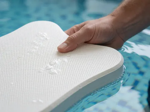 Main adulte tenant un panneau de natation blanc texturé partiellement immergé dans l'eau claire avec des bulles sur des carreaux bleus de piscine.