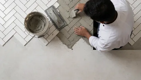 Un carreleur applique du joint gris sur des carreaux blancs en motif chevrons, avec une spatule et un seau à proximité.