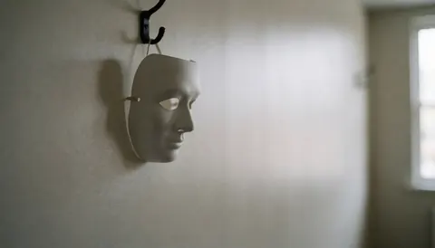 Masque de théâtre blanc accroché à un crochet sur un mur texturé, éclairage tamisé et arrière-plan flou, idéal pour un atelier de théâtre.