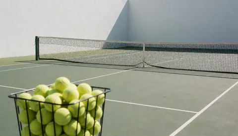 Terrain de tennis extérieur avec un panier rempli de balles jaunes, un filet et des murs blancs, idéal pour un site web d'instructeur de tennis.