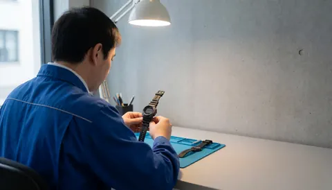 Horloger travaillant sur une montre numérique posée sur une grille bleue, avec un mur en béton en arrière-plan et un espace réservé au texte.