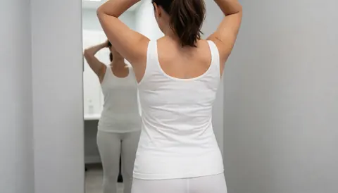 Vue de dos d'une femme ajustant ses cheveux devant un miroir dans un vestiaire aux murs gris clair, idéal pour une école de danse.
