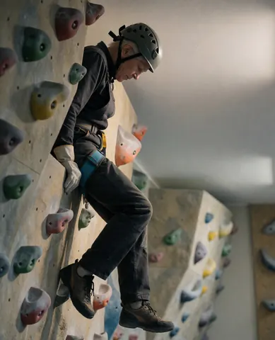 Un client senior portant casque et harnais, posant entre les prises sur un mur d'escalade artificiel dans un gymnase éclairé doucement.