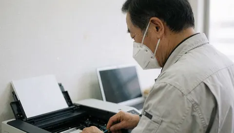 Technicien portant un masque N95 effectuant une réparation sur une imprimante dans un atelier propre avec un ordinateur portable, illustrant un service professionnel.