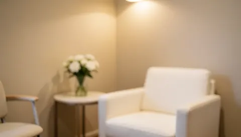 Salle d'attente avec des chaises blanches, une petite table avec des fleurs et des murs beige, idéale pour les salons de beauté ou les spas.