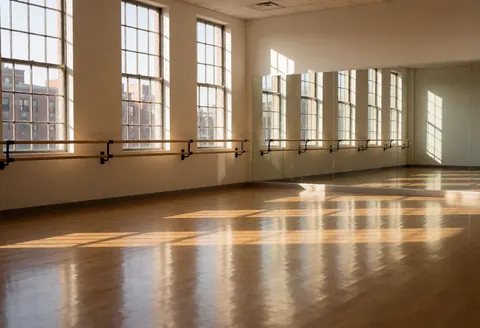 Studio de danse vide avec sol en bois poli, barre de ballet et miroirs, éclairé par la lumière naturelle de fenêtres hautes, idéal pour les cours de danse.