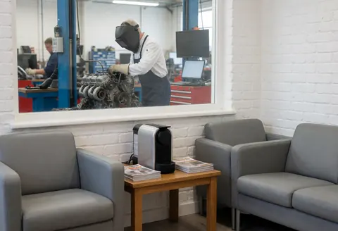 Salle d'attente avec chaises grises et cafetière, un mécanicien répare un moteur visible par une fenêtre en verre.