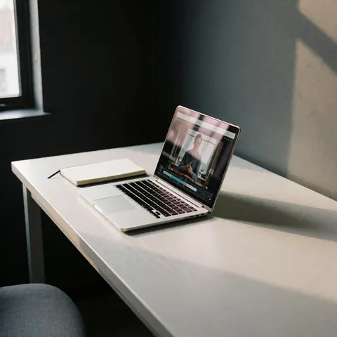 Ordinateur portable ouvert montrant un cours de programmation, accompagné d'un cahier, posé sur un bureau blanc dans un bureau aux murs gris et à la lumière naturelle.
