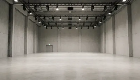 Salle d'événements vide avec murs en béton, plafonds à structures métalliques et éclairage professionnel, idéal pour événements et présentations.
