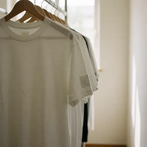 T-shirt blanc avec patch brodé sur la manche, suspendu à un cintre en bois dans un vestiaire aux murs blancs et à la lumière naturelle.
