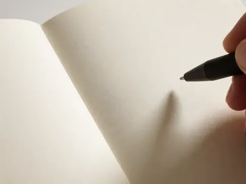 Main tenant un stylo noir au-dessus d'un carnet ouvert sur un papier à texture visible, éclairage chaleureux, idéal pour la photographie de produit.