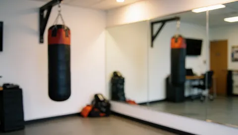 Fond de studio de boxe avec sac noir et rouge, grand miroir et murs blancs, éclairage chaleureux adapté pour textes.