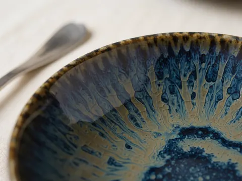 Détail d'un bol en céramique artisanale avec motif craquelé bleu et doré posé sur une nappe crème, idéal pour la décoration et la mise en scène de banquets.