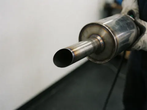 Silencieux d'échappement automobile tenu par des mains gantées de cuir, montrant des soudures dans un atelier au sol sombre et mur blanc.