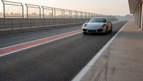 Voiture de sport argentée stationnée sur une piste de course au lever du jour, avec barrières de sécurité et marquages au sol visibles.