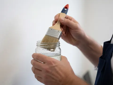 Gros plan d'une main tenant un pinceau au-dessus d'un récipient contenant du solvant, sur un fond blanc uni, illustrant le nettoyage des outils de peinture.