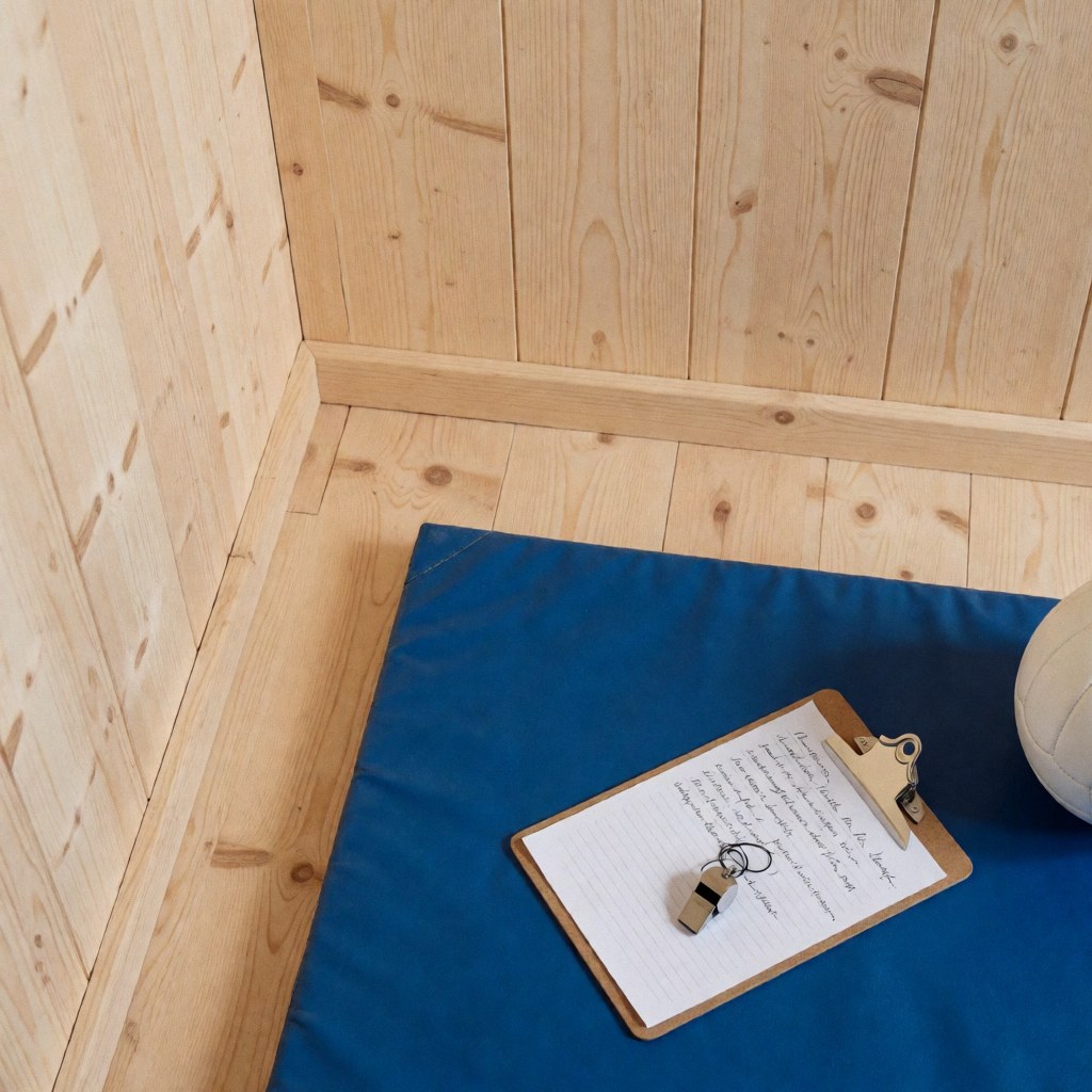 Portefeuille d'entraîneur de volleyball posé sur une surface avec un tableau tactique, un sifflet et un ballon de volleyball sur un sol bleu et des murs en bois.