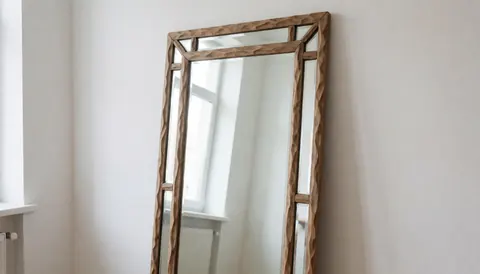 Miroir artisanal avec un cadre en bois sculpté, reflétant la lumière naturelle et un mur blanc dans un atelier de verrerie.