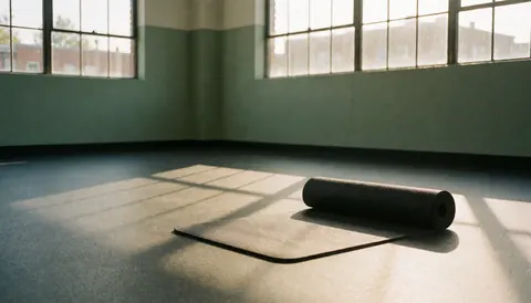 Tapis de yoga noir enroulé sur un sol en caoutchouc texturé, éclairé par la lumière du matin entrant par des fenêtres industrielles dans une salle de sport vide.