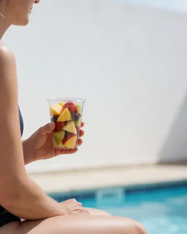 Une femme profite d'un fruit frais au bord d'une piscine, avec un mur blanc et de l'eau bleue sous un soleil éclatant, idéal pour une entreprise de piscines publiques.