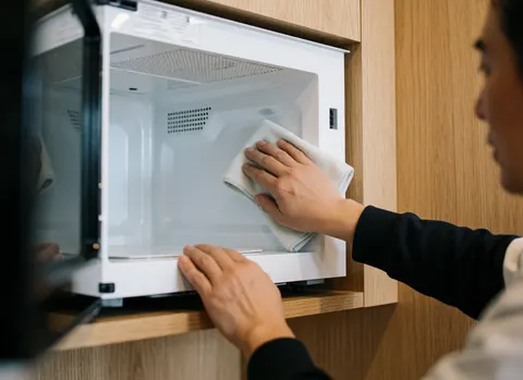 Une main nettoie l'intérieur brillant d'un micro-ondes blanc avec une serviette en microfibre dans une cuisine aux murs en bois clair.