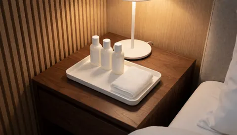Plateau blanc contenant des produits de toilette et une serviette posés sur une table de nuit en bois, avec un lit aux draps blancs et un mur à lamelles en arrière-plan.