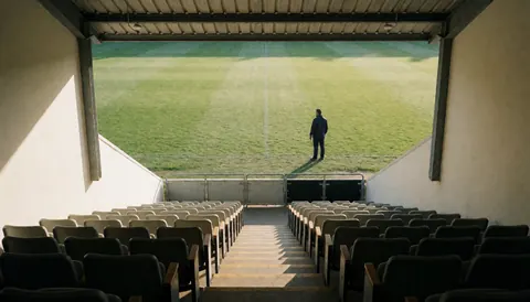 Entraîneur de football debout sur le terrain, vu depuis les gradins vides, sous la lumière du matin, idéal pour la page d'accueil d'un site web.