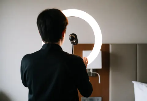 Un maquilleur professionnel règle un anneau lumineux dans une chambre d'hôtel avec tête de lit et murs neutres, préparant son équipement pour un événement.