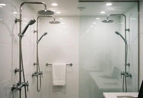 Deux cabines de douche en verre avec robinetterie inox, murs blancs, serviette blanche suspendue, idéal pour les centres de bien-être.