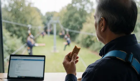Un homme portant un harnais bleu observe le tyrolienne depuis une fenêtre tout en tenant une barre énergétique, avec un ordinateur portable sur la table, idéal pour une page d'accueil de parc d'aventure.