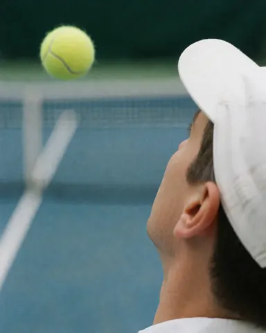 Un joueur de tennis portant une casquette blanche observe la balle en vol. Le filet est visible sur un court bleu, idéal pour illustrer des services d'entraînement.