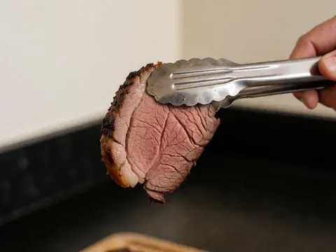 Morceau de viande grillée tenu par des pincettes métalliques sur un fond de cuisine sombre avec des murs blancs chauds et une texture de graisse brillante.