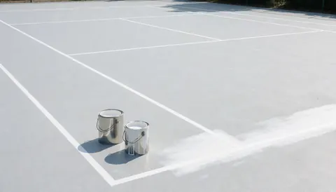 Deux contenants de peinture blanche posés sur la surface d'un court de tennis avec des lignes fraîchement tracées, illustrant les travaux d'entretien ou d'installation.