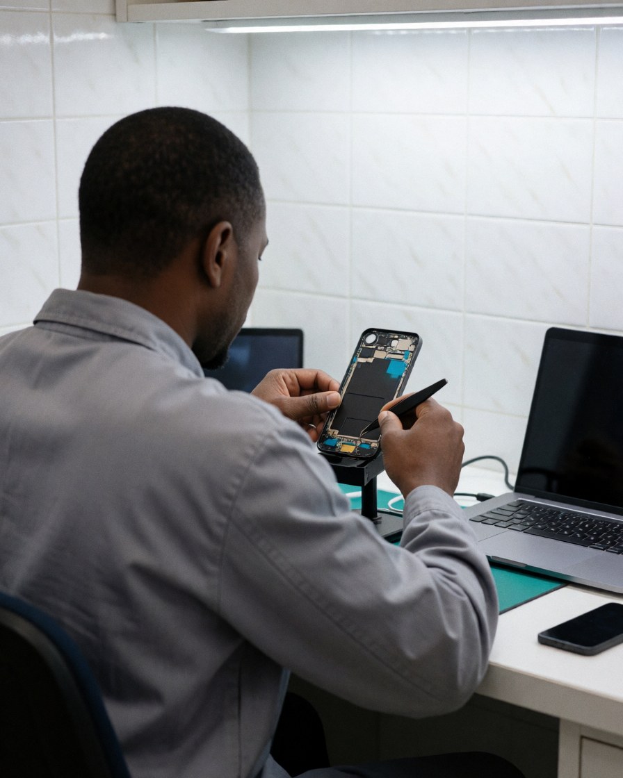 Un technicien utilise des pinces pour réparer un téléphone démonté dans un atelier aux murs carrelés blanc avec un ordinateur portable.