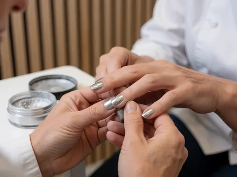 Détail d'une manucure professionnelle avec poudre métallique brillante sur les ongles, outils et fond en bois clair visibles