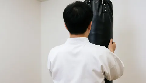 Un pratiquant de karate en tenue blanche s'entraîne sur un sac de frappe noir dans un dojo aux murs blancs, illustrant un service en action.