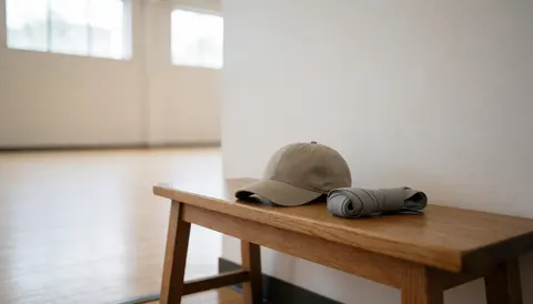 Un chapeau de baseball et des gants de boxe gris posés sur un banc en bois à l'intérieur d'un dojo aux murs blancs et aux fenêtres haisses.