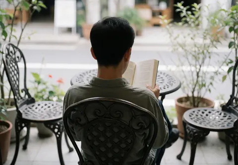 Un homme détendu lit un livre assis sur une chaise en fer forgé dans une terrasse extérieure verdoyante, ambiance calme et reposante.