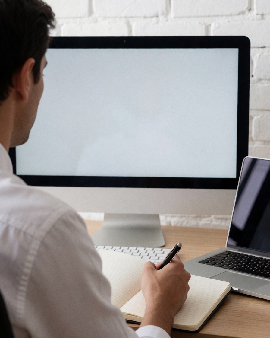 Un professionnel note des informations dans un carnet tout en consultant son écran d'ordinateur dans un bureau lumineux avec un mur en briques blanches.