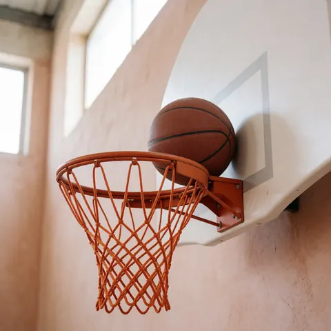 Un ballon de basket repose sur le bord d'un panier de basketball dans un gymnase aux murs en terre cuite baigné de lumière naturelle.