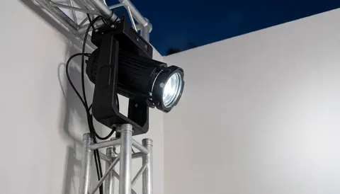 Projecteur professionnel fixé à une structure en treillis pointant vers un sol vide, avec des murs blancs et un ciel bleu foncé en arrière-plan.