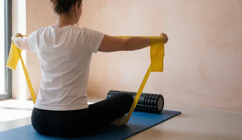Une femme adulte effectue des étirements avec une bande de résistance jaune sur un tapis bleu, près d'un rouleau de mousse noir, dans un studio de gymnastique aux murs terracotta.