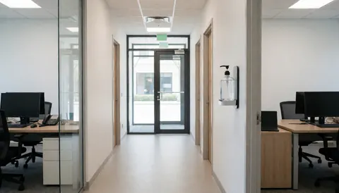 Couloir de bureau moderne avec distributeur de gel désinfectant mural, bureaux avec ordinateurs et portes vitrées, idéal pour la propreté professionnelle.